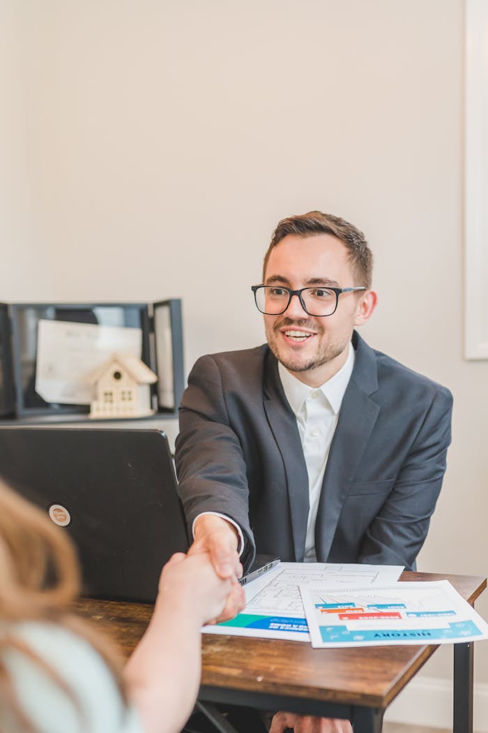 A professional real estate agent welcomes a client in the office with documents on the desk.